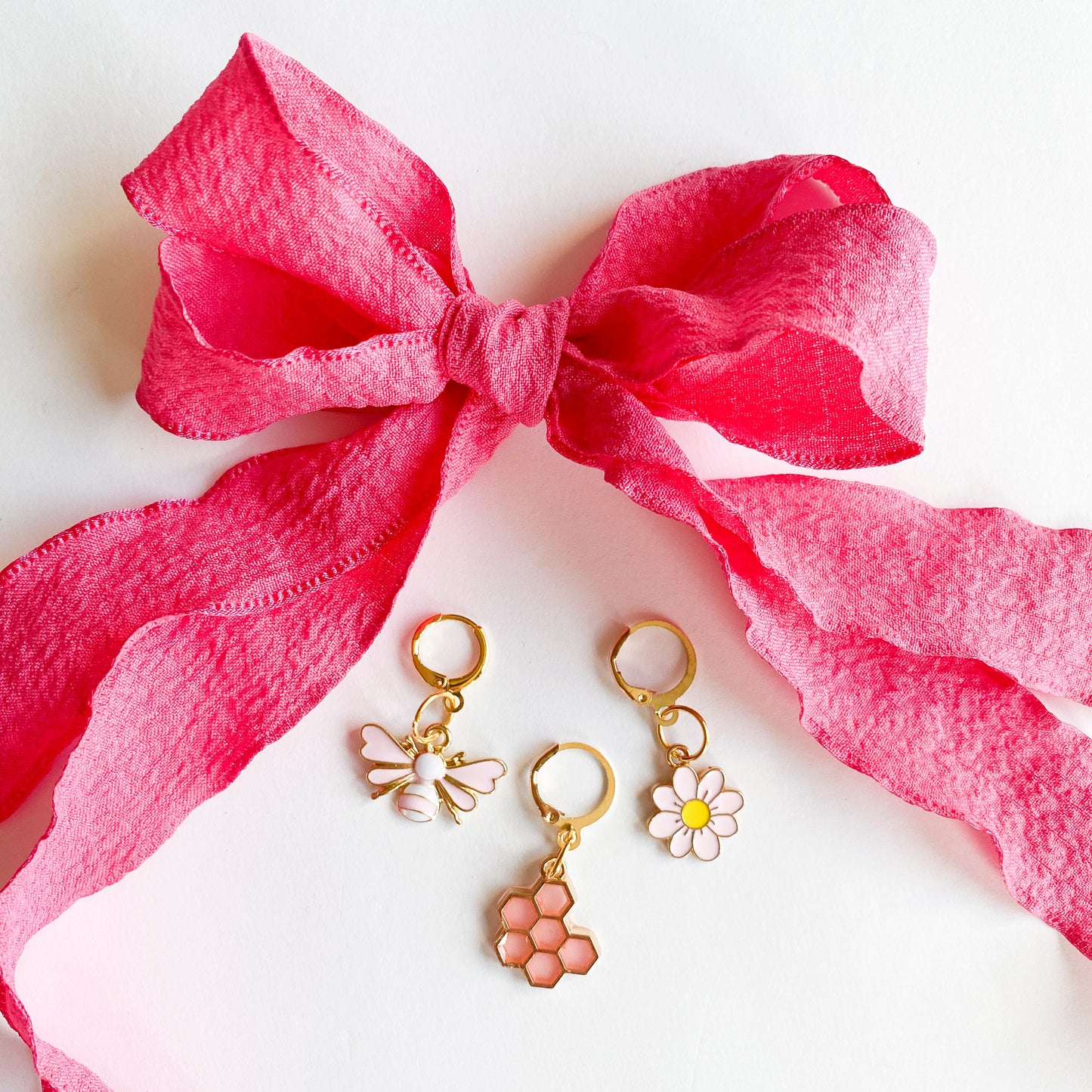 Gold shoe charms with pink bee, honeycomb, and white daisy designs, displayed with bright pink ribbon shoelaces tied in a bow on white background.