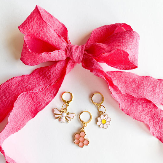 Gold shoe charms with pink bee, honeycomb, and white daisy designs, displayed with bright pink ribbon shoelaces tied in a bow on white background.