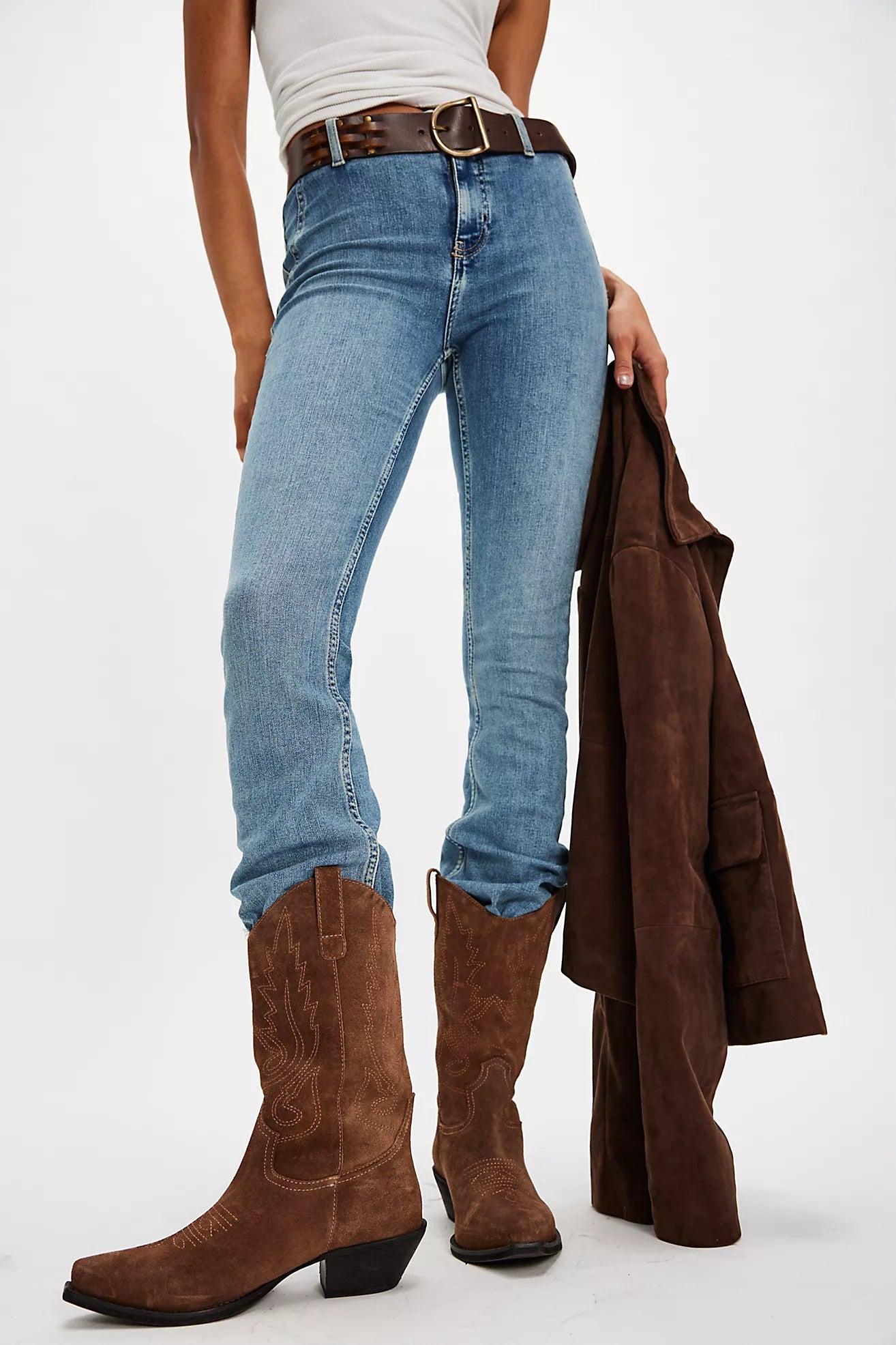 Person wearing blue jeans and brown boots holding a brown jacket on a white background