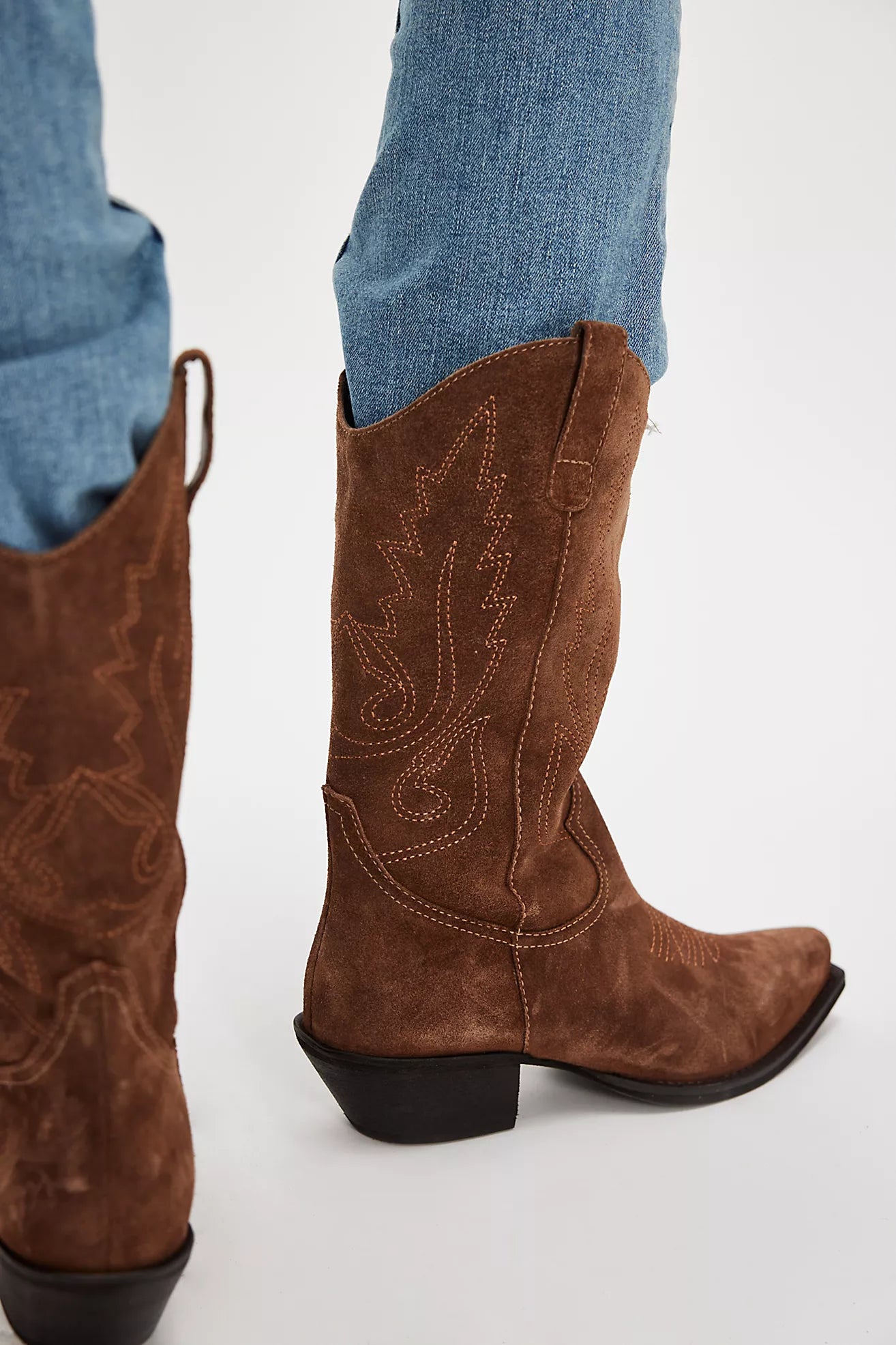 Brown cowboy boots with intricate designs worn with blue jeans on a white background