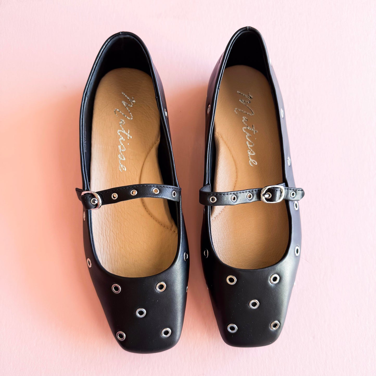 Top view of red and black studded Mary Jane ballet flats with metal grommets, placed on a soft pink background.