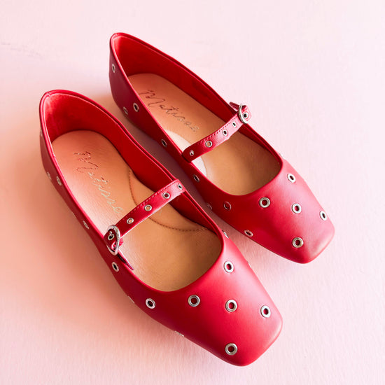 Top view of red  studded Mary Jane ballet flats with metal grommets, placed on a soft pink background.