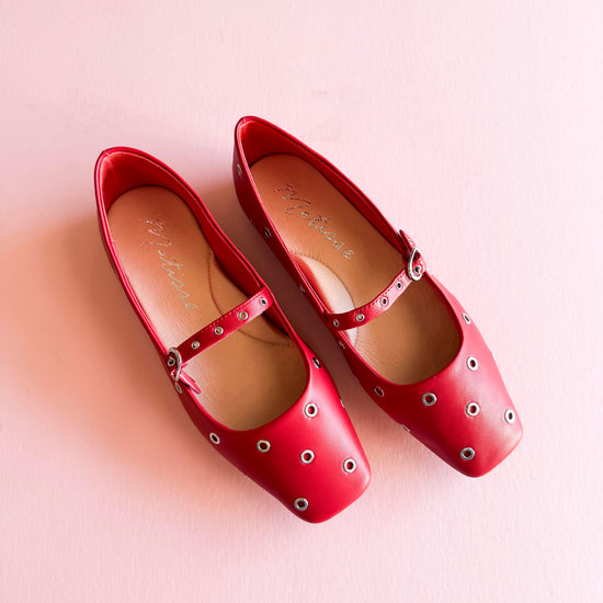 Top view of red  studded Mary Jane ballet flats with metal grommets, placed on a soft pink background.