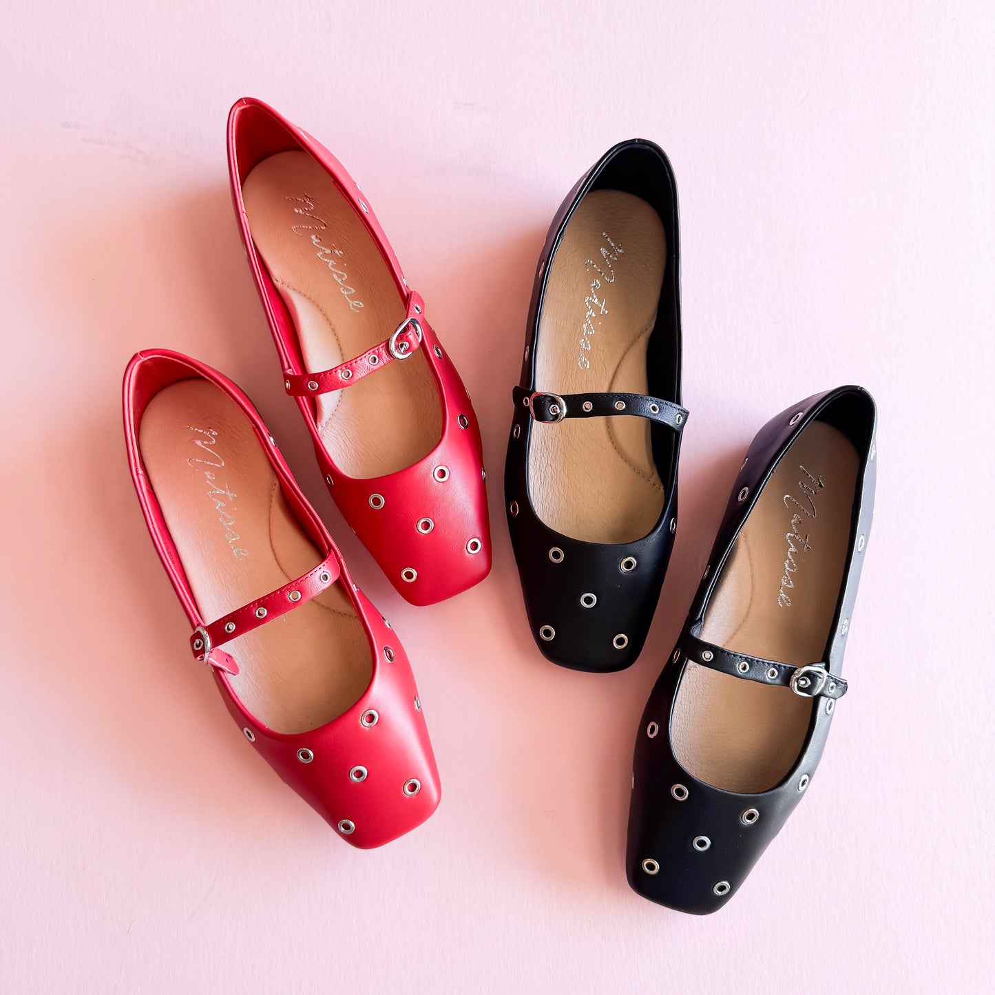 Top view of red and black studded Mary Jane ballet flats with metal grommets, placed on a soft pink background.