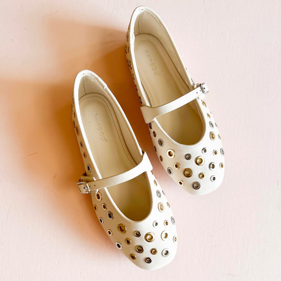 Cream ivory studded ballet flats with gold and silver metal eyelet accents, featuring an adjustable strap and rounded toe design, displayed on a light pink background.