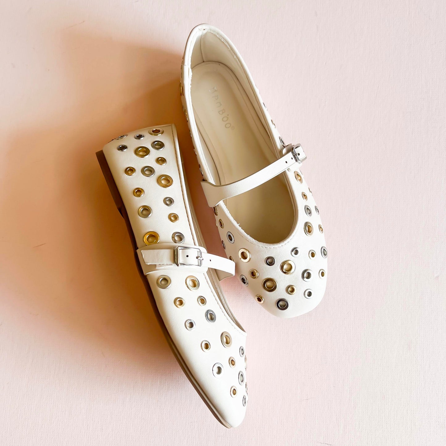 Cream ivory studded ballet flats with gold and silver metal eyelet accents, featuring an adjustable strap and rounded toe design, displayed on a light pink background.