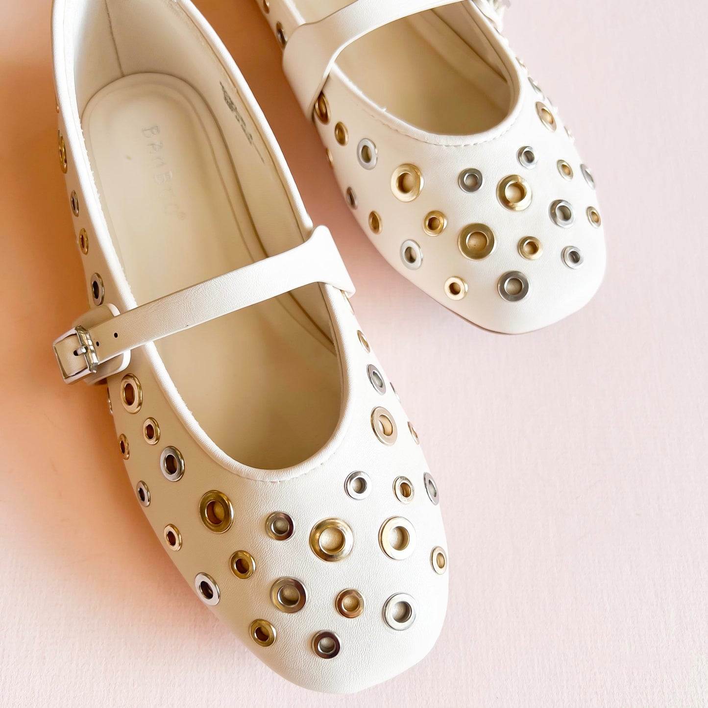 Cream ivory studded ballet flats with gold and silver metal eyelet accents, featuring an adjustable strap and rounded toe design, displayed on a light pink background.