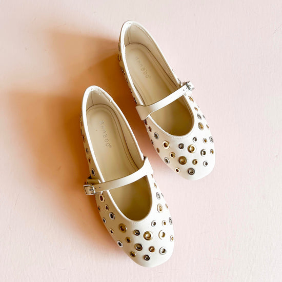 Cream ivory studded ballet flats with gold and silver metal eyelet accents, featuring an adjustable strap and rounded toe design, displayed on a light pink background.