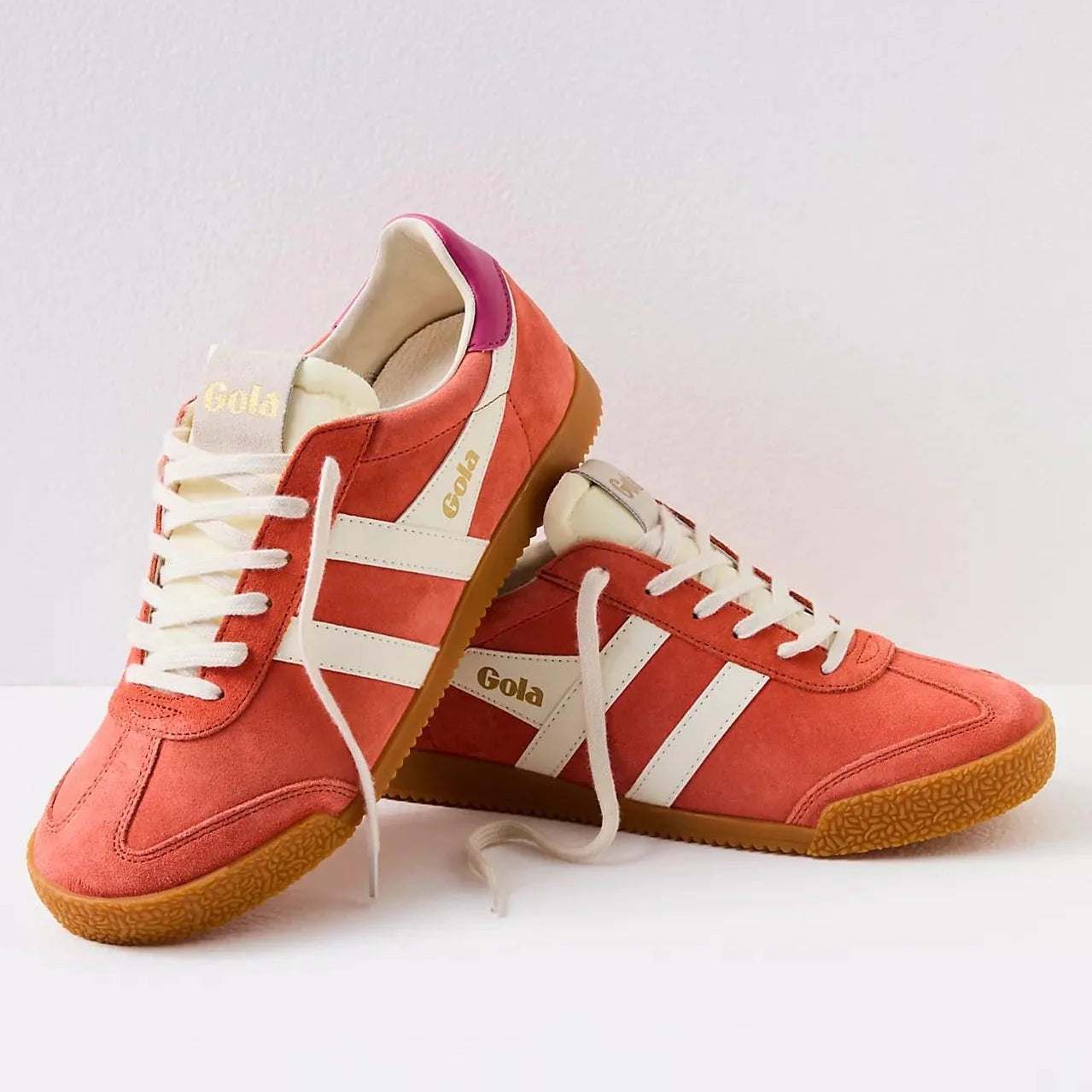 Gola sneakers in a vibrant color scheme of hot coral, off-white, and fuchsia. The shoes have a classic retro design with suede material and a gum rubber sole. The brand name "Gola" is prominently displayed on the side and tongue of the sneakers in gold lettering. The laces and stripes are off-white, providing a striking contrast to the bold coral and fuchsia colors.