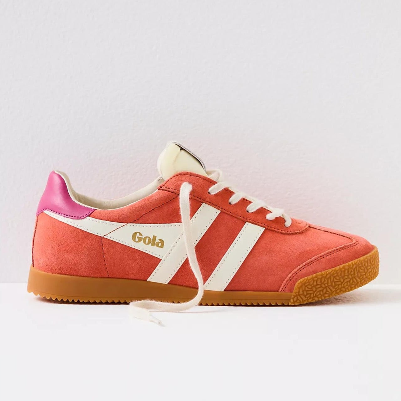 Gola sneakers in a vibrant color scheme of hot coral, off-white, and fuchsia. The shoes have a classic retro design with suede material and a gum rubber sole. The brand name "Gola" is prominently displayed on the side and tongue of the sneakers in gold lettering. The laces and stripes are off-white, providing a striking contrast to the bold coral and fuchsia colors.