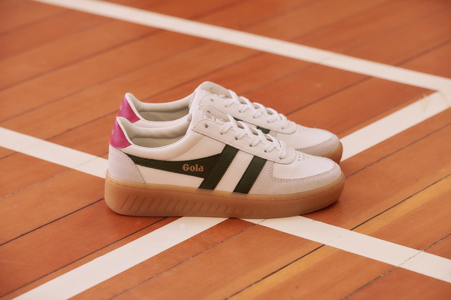 White and green retro-style sneakers by Gola, The shoes feature a white upper with green stripes and a pink heel tab, contrasted against a gum sole.