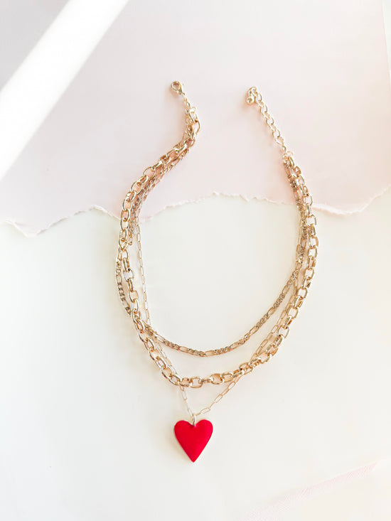 A stylish layered gold chain necklace featuring a bright red heart pendant, set against a softly lit, textured white background with torn paper detail