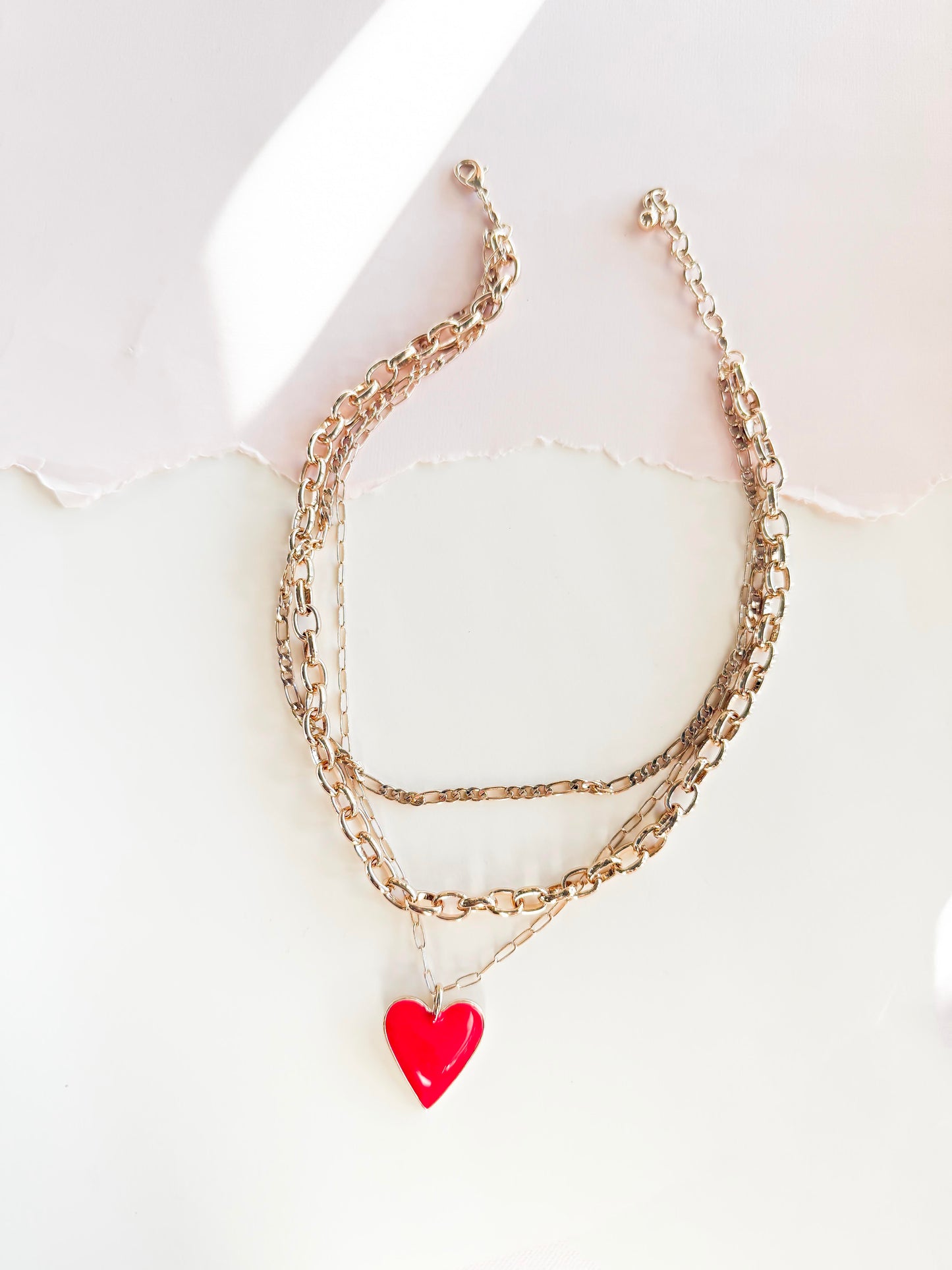 A stylish layered gold chain necklace featuring a bright red heart pendant, set against a softly lit, textured white background with torn paper detail