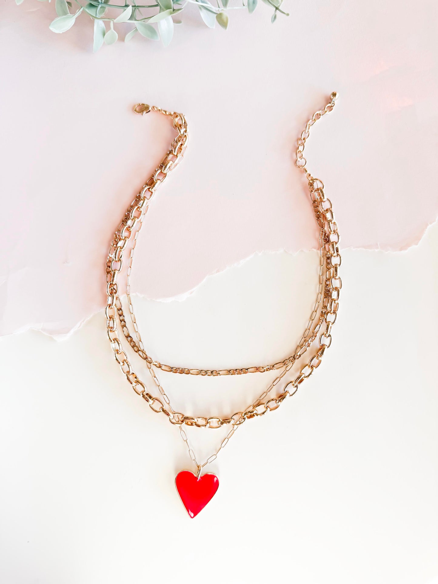 A stylish layered gold chain necklace featuring a bright red heart pendant, set against a softly lit, textured white background with torn paper detail