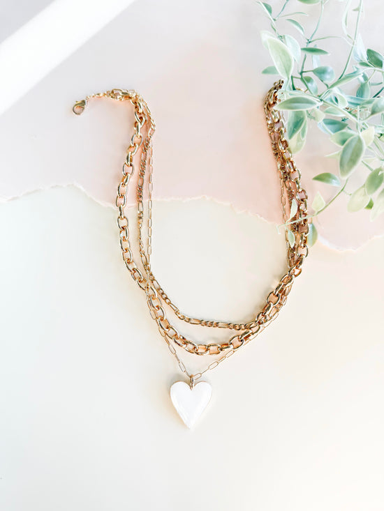 A stylish layered gold chain necklace featuring a bright white heart pendant, set against a softly lit, textured white background with torn paper detail