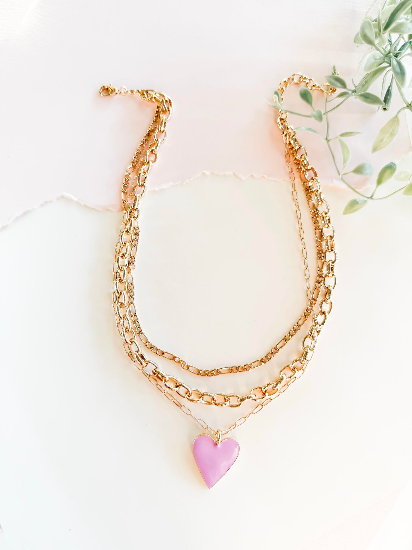 A stylish layered gold chain necklace featuring a bright lavender heart pendant, set against a softly lit, textured white background with torn paper detail