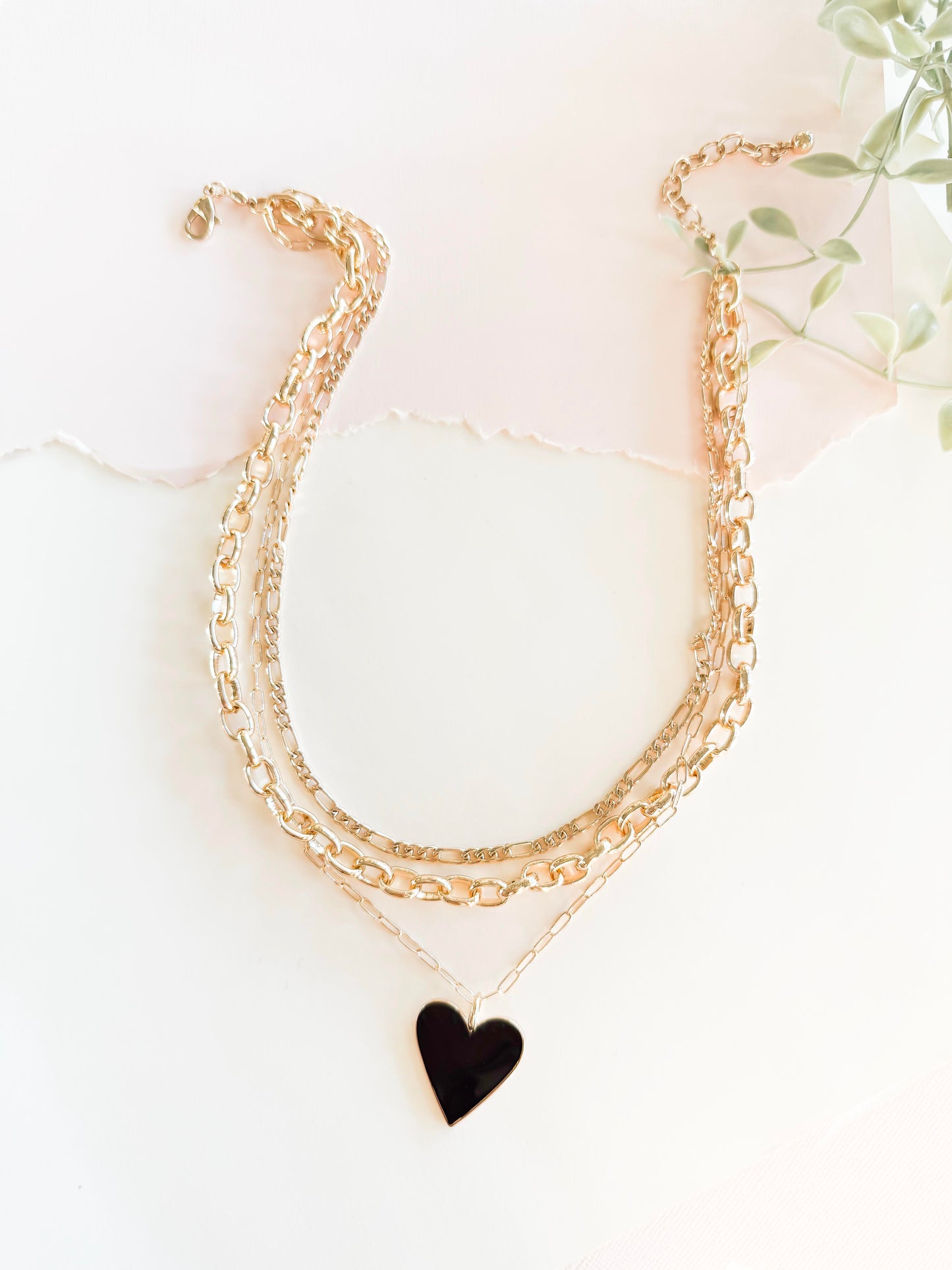 A stylish layered gold chain necklace featuring a bright black heart pendant, set against a softly lit, textured white background with torn paper detail