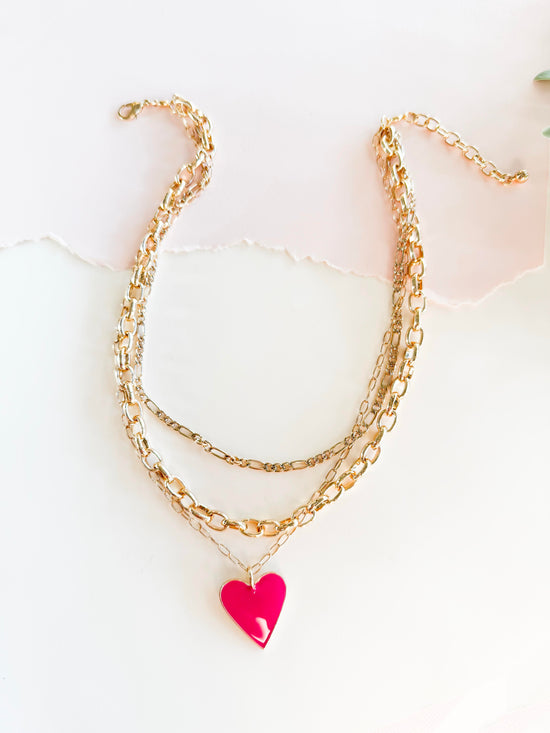 A stylish layered gold chain necklace featuring a bright heart pendant, set against a softly lit, textured white background with torn paper detail