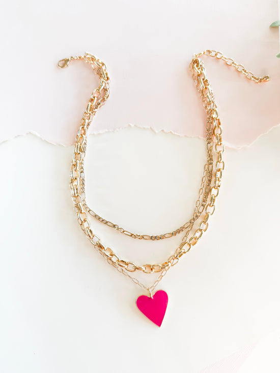 A stylish layered gold chain necklace featuring a bright pink heart pendant, set against a softly lit, textured white background with torn paper detail