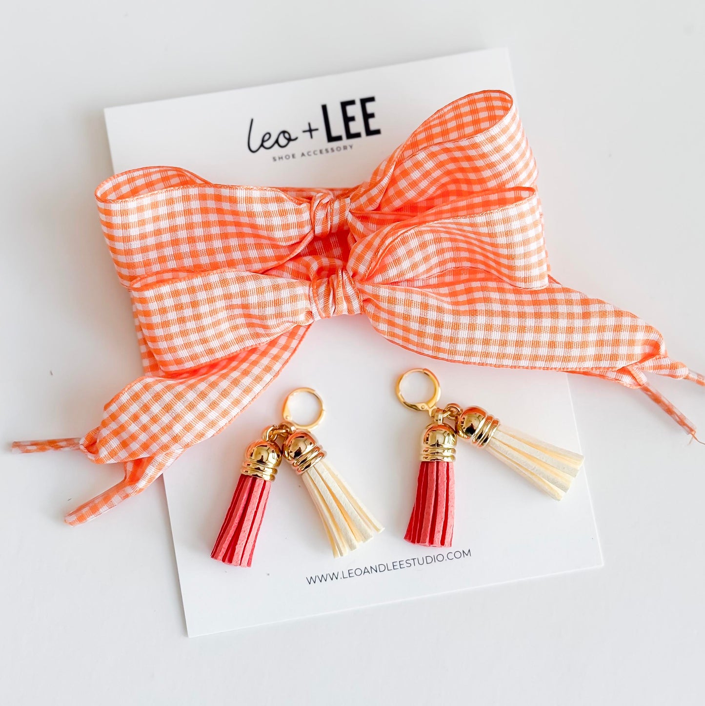 Two pink and cream  tassel charms with gold accents on a card featuring 'leo + LEE' brand name.