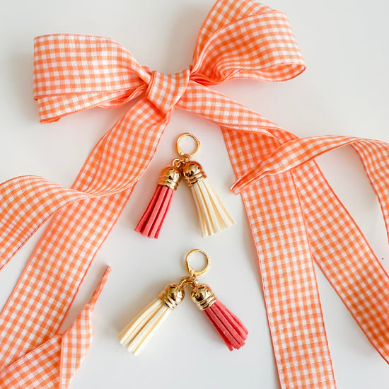Two tassel charms with gold accents on a white background with an orange checkered ribbon.