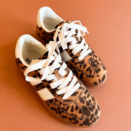 Leopard-print sneakers with white laces and contrasting white stripes, displayed on a warm terracotta background.