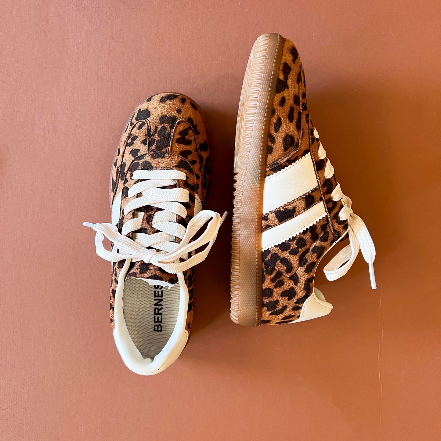 Leopard-print sneakers with white laces and contrasting white stripes, displayed on a warm terracotta background.