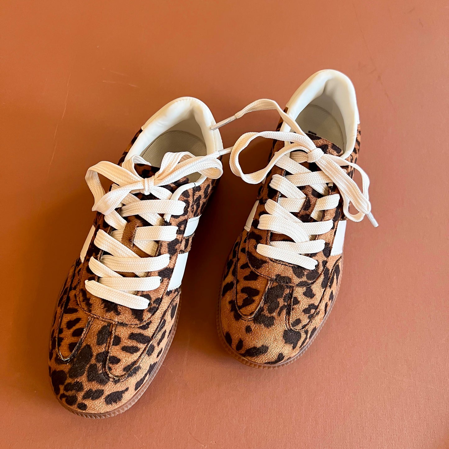 Leopard-print sneakers with white laces and contrasting white stripes, displayed on a warm terracotta background.