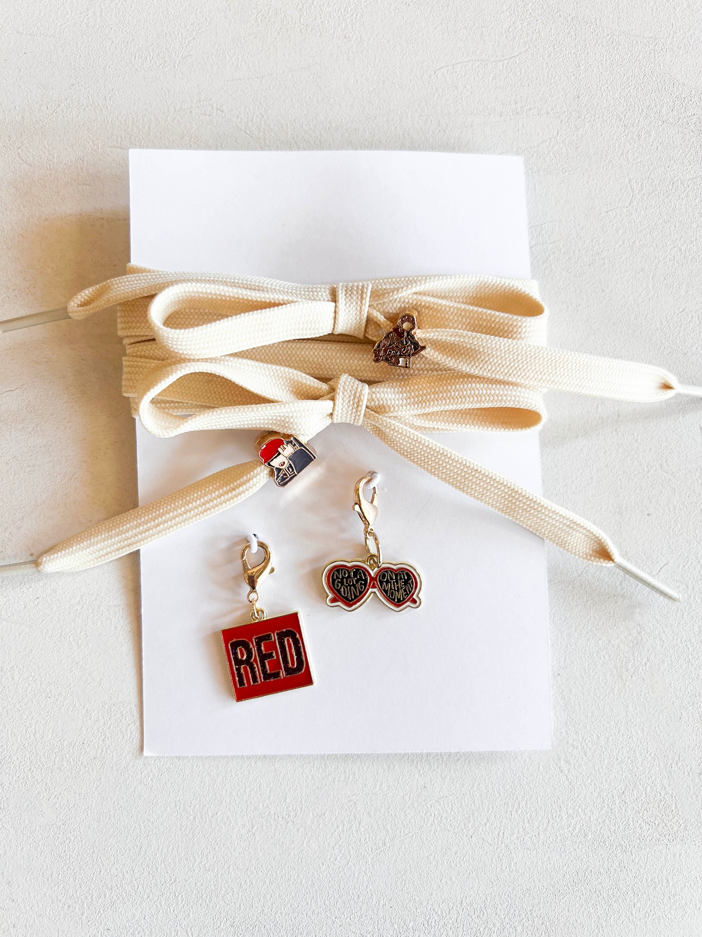 Sneaker with custom Swiftie-themed charms on cream-colored laces, featuring a 'RED' square charm, heart-shaped sunglasses charm, and two additional playful designs displayed next to extra laces.