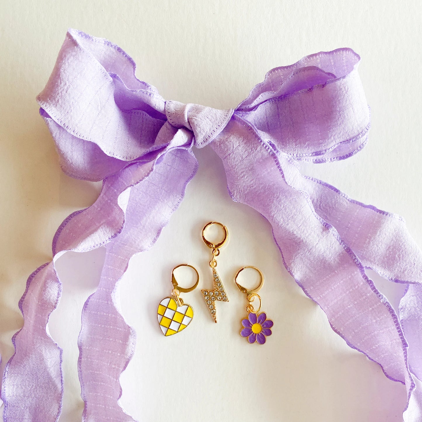 Gold shoe charms with yellow checkered heart, rhinestone lightning bolt, and purple flower, displayed with large lavender ribbon shoelaces tied in a bow on white background.