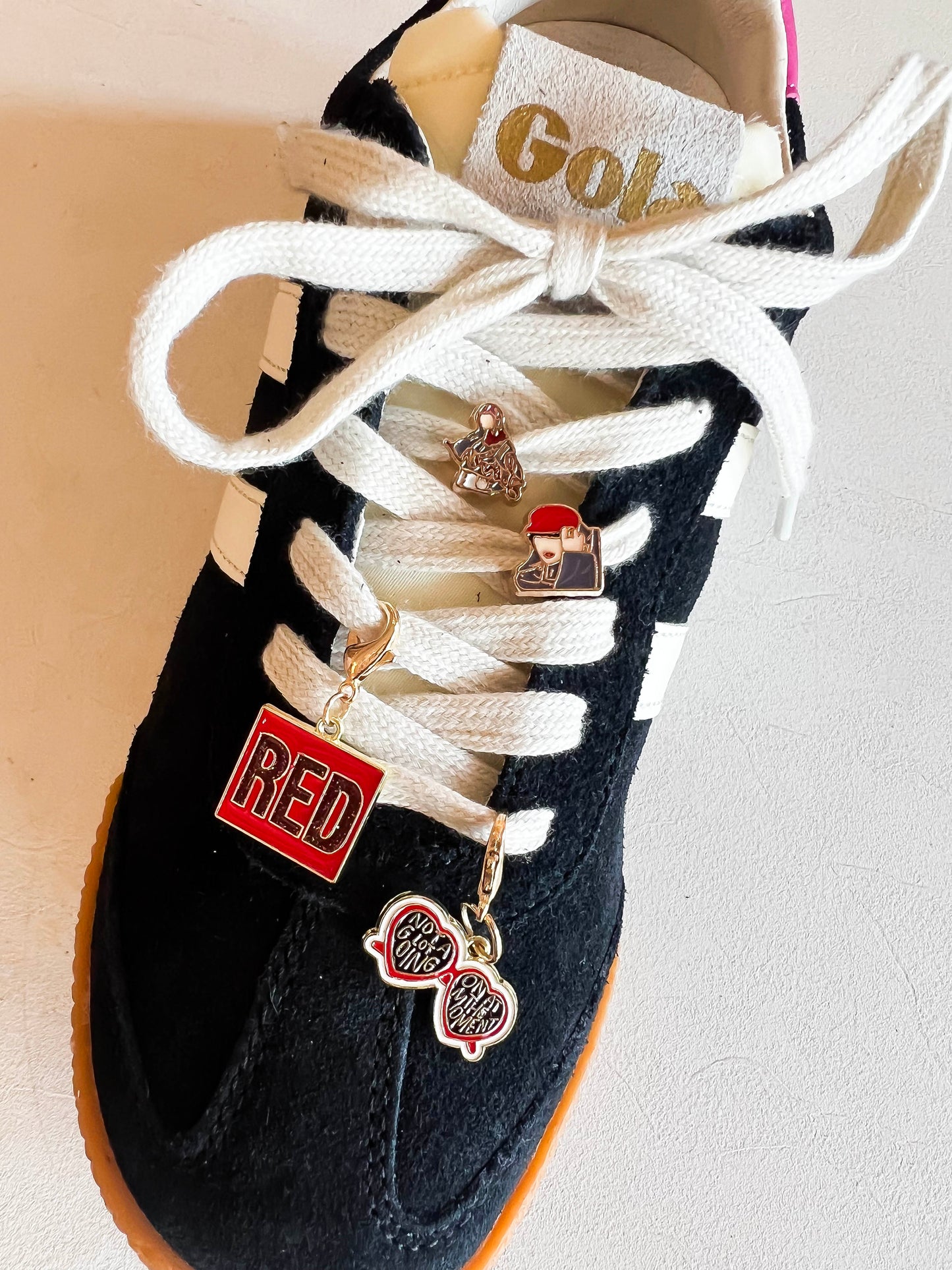 Sneaker with custom Swiftie-themed charms on cream-colored laces, featuring a 'RED' square charm, heart-shaped sunglasses charm, and two additional playful designs displayed next to extra laces.