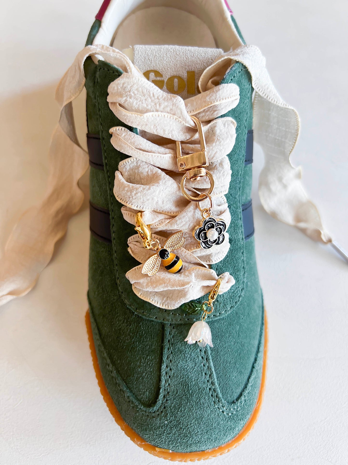 Green sneaker with decorative lace featuring bee and flower charms, cream-colored fabric laces, and a gold keychain attachment