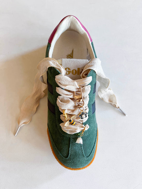 Green sneaker with decorative lace featuring bee and flower charms, cream-colored fabric laces, and a gold keychain attachment